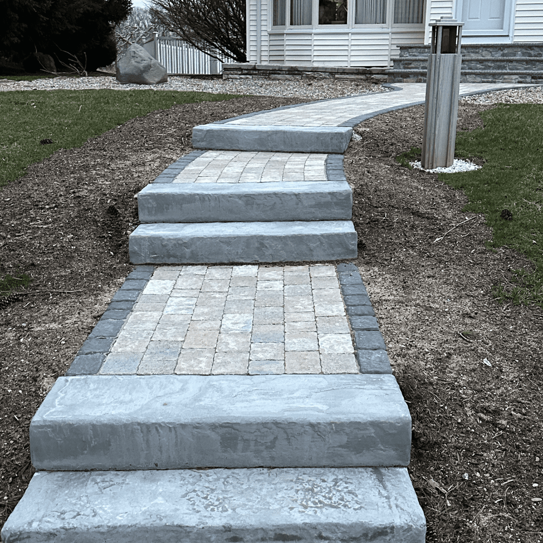 Winding paved walkway with stone steps leading up to a cozy white house with a large front window and a lamp post on the side.