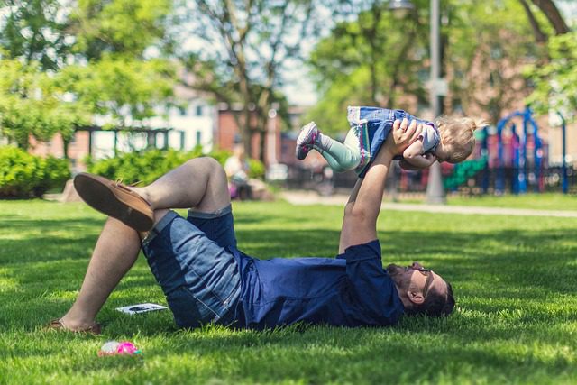 father holding his child above him while laying on lush green grass.